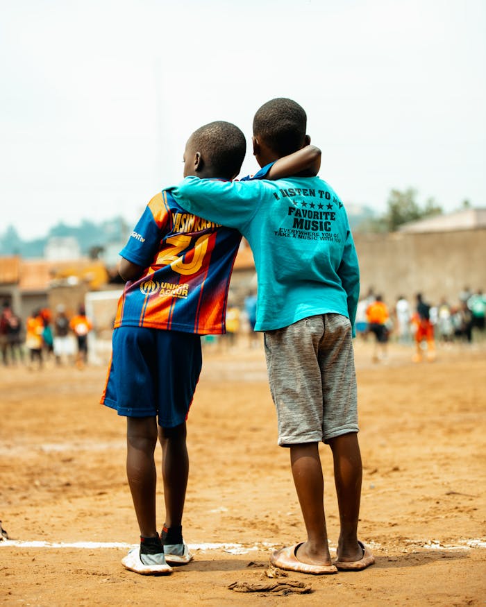 Two young boys embrace on a dirt soccer field in Kampala, Uganda. Friendship and sports unite.